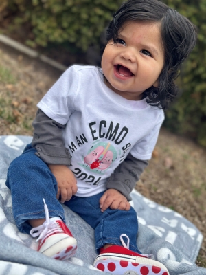 A smiling baby with curly black hair sits on a grey blanket outside. He is wearing a t-shirt that reads “I am an ECMO survivor – 2024."