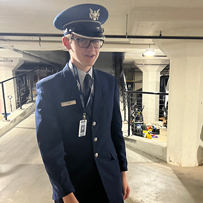 A tall teen boy poses in a parking garage. He is wearing a navy blue formal military uniform, including a suit jacket, creased pants and a white dress shirt and blue tie. He is wearing a blue cap with a black brim and a gold eagle insignia.