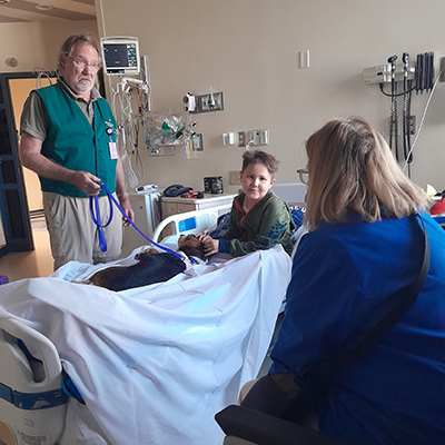 Jaxson sits in a hospital bed interacting with a therapy animal while two adults visit in the patient room.