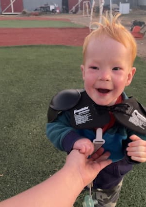 Young child wearing a supportive harness while standing on an outdoor athletic field and holding an adult’s hand.
