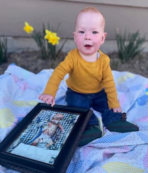 Infant sitting outdoors on a blanket and holding a framed photograph of another infant