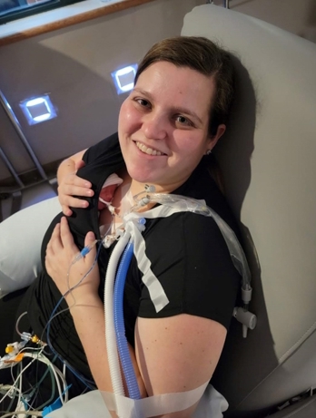 Person seated in a hospital chair holding an infant while connected to medical monitoring equipment and respiratory tubing.