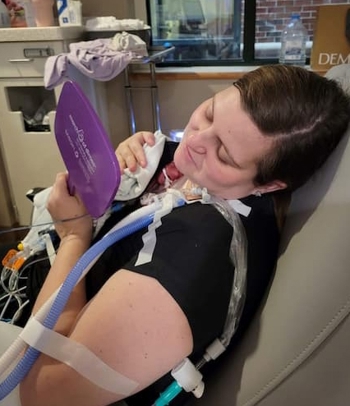 Person seated in a hospital chair holding an infant while connected to medical equipment.