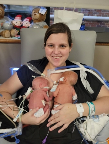 Adult seated in a hospital chair holding two infants receiving medical care, with monitoring leads, tubing and clinical equipment visible