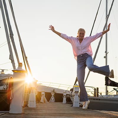A smiling young woman is leaping in the air. She’s on a dock with sailboats and the setting sun behind her. She’s wearing jeans, a pink button-down shirt, and white tennis shoes. 