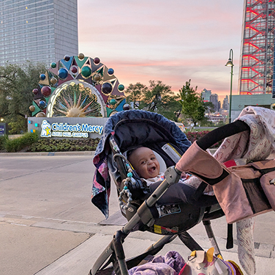 Lilly sits in a stroller outside Children's Mercy Adele Hall Campus with hospital signage visible in the background.