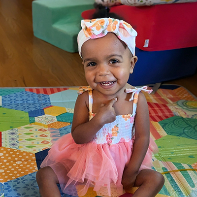 Lilly sits on a colorful play mat wearing a patterned dress and headband.