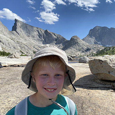 Child wearing a sun hat standing outdoors in a rocky mountain landscape, with large granite peaks and a blue sky in the background