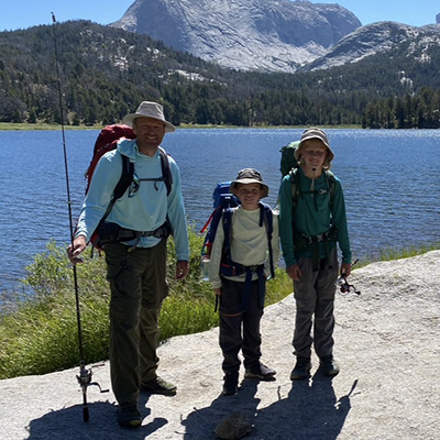Three hikers wearing backpacks standing beside a mountain lake, with forested slopes and granite peaks in the background.