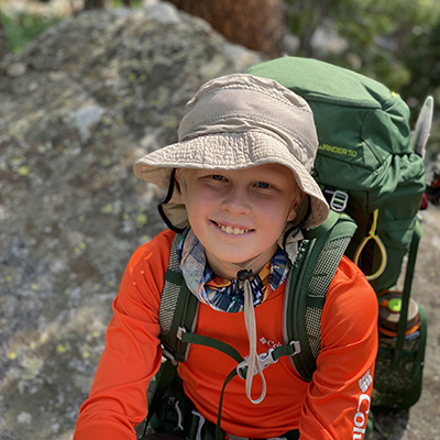 Young person wearing a sun hat and orange hiking shirt with a large green backpack in a rocky, wooded outdoor setting.