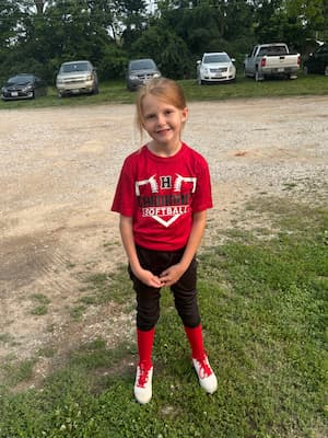 A young girl smiles wearing a red, black and white softball uniform in a gravel parking lot. Her shirt reads “H Cardinals Softball.”  