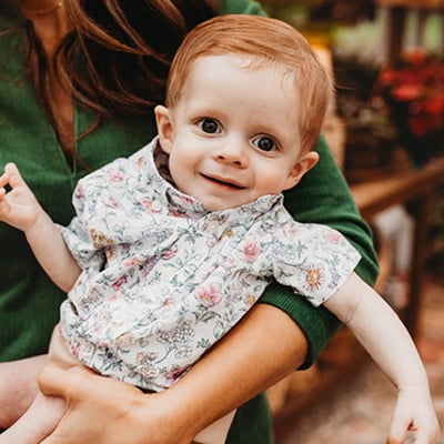 Infant in a floral outfit being held by an adult, with an indoor setting visible in the background.