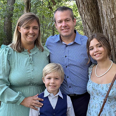 A family of four (a mom, dad, teenage daughter and grade-school aged boy) poses for a portrait with trees in the background. They are wearing dress clothes in pastel greens and blues.