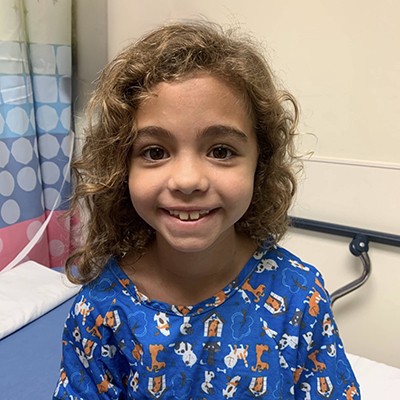 A young girl grins at the camera while sitting at the edge of a hospital bed. She has wavy light brown hair and is wearing a blue hospital gown with cartoon dogs and cats on it. 
