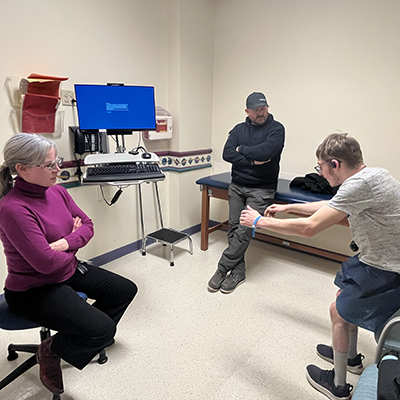 A doctor, teen patient and father are in a medical exam room. The teen, in a gray shirt and blue shorts, stands in front of a chair with his arms outstretched. His doctor, a woman in a purple turtleneck, sits on a stool and observes him. His father leans against the exam table with his arms and legs crossed watching his son.