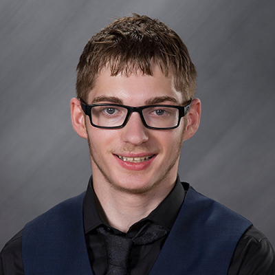 12th grade school photo of a young man with brown hair, black glasses and a slight beard. He wears a black dress shirt and black tie with a navy blue vest over it.