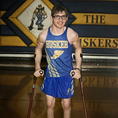 A muscular teen athlete stands in a gym with a picture of the school mascot and sign reading “Home of the Huskers” behind him. He is wearing a blue and gold track tank top and blue shorts and uses arm crutches for support.