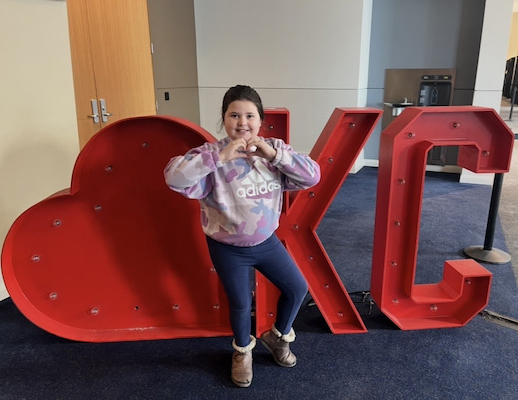 Child standing in front of large red KC letters and making a heart shape with their hands.