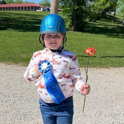 Child wearing a blue riding helmet holding a blue award ribbon and an orange flower outdoors.
