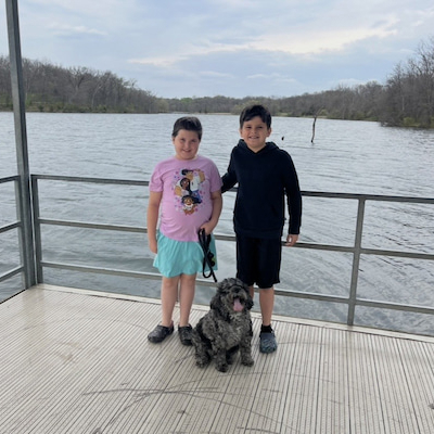 Two children standing with a dog on a dock overlooking a lake.
