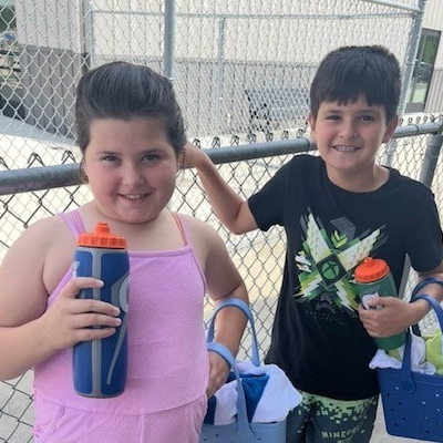 Two children standing side by side outdoors near a chain-link fence, holding reusable water bottles and carrying tote bags.