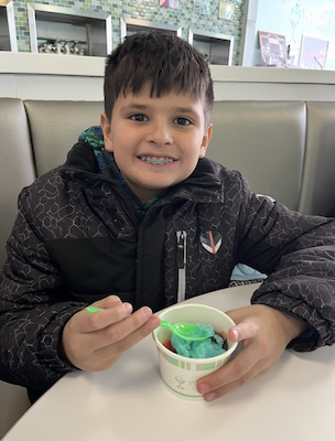 Child with braces holding a cup of green frozen dessert while seated at a table.