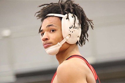 Student athlete wearing wrestling headgear and a singlet, standing on a gymnasium floor during a wrestling event.