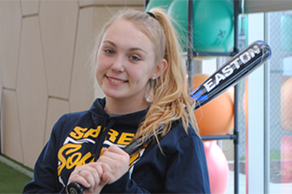 Student athlete holding a softball bat while standing in an indoor rehabilitation or sports performance facility, with exercise equipment visible in the background.
