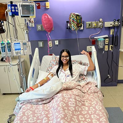 Patient resting in a hospital bed with IV tubing and medical monitoring equipment, raising one arm.