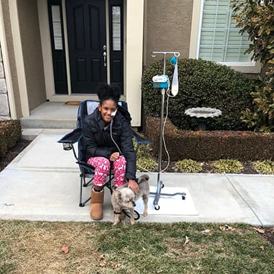 Person seated outside a home beside an IV pole, with a small dog standing nearby.