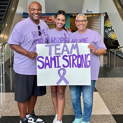 Three people wearing purple shirts and holding a handmade sign that reads 'TEAM SAMI STRONG' with a purple awareness ribbon.