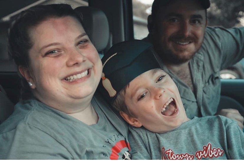 Two adults and a child seated together inside a vehicle, with the child wearing a graduation cap