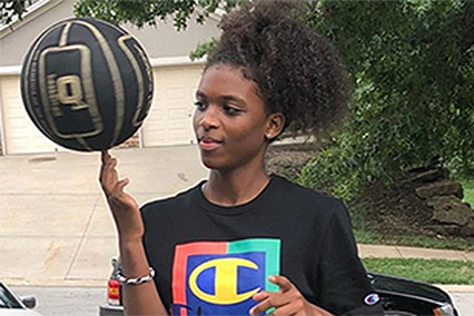 Young athlete balancing a spinning basketball on one finger while standing outside near a driveway and trees.