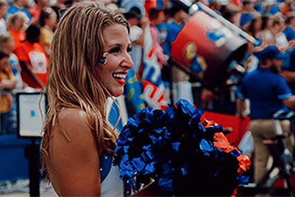 Cheerleader holding blue and orange pom-poms at a stadium event, with a crowd and sports-related activities visible behind them.