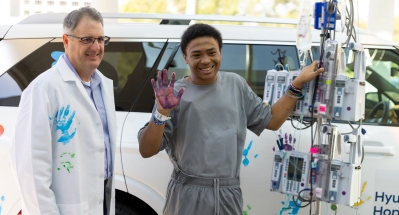 Patient standing beside a medical infusion pole outdoors, raising a hand covered in purple paint next to a clinician in a white coat with a blue handprint. A white vehicle is visible in the background.