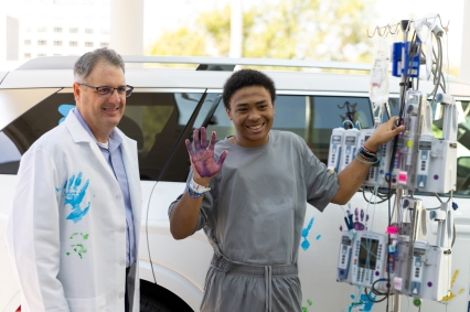 Patient standing beside a medical infusion pole outdoors, raising a hand covered in purple paint next to a clinician in a white coat with a blue handprint. A white vehicle is visible in the background.
