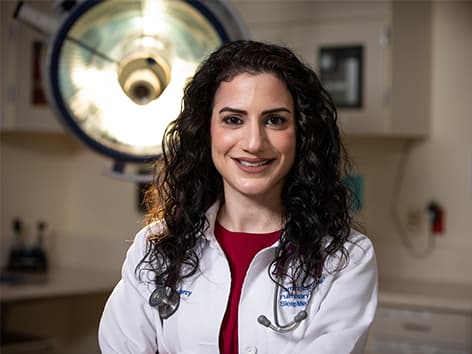 Female clinician in white coat, brown hair with medical equipment in the background.  