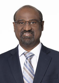 A man with a goatee and glasses is wearing a white, collared shirt and tie with a dark blue blazer.
