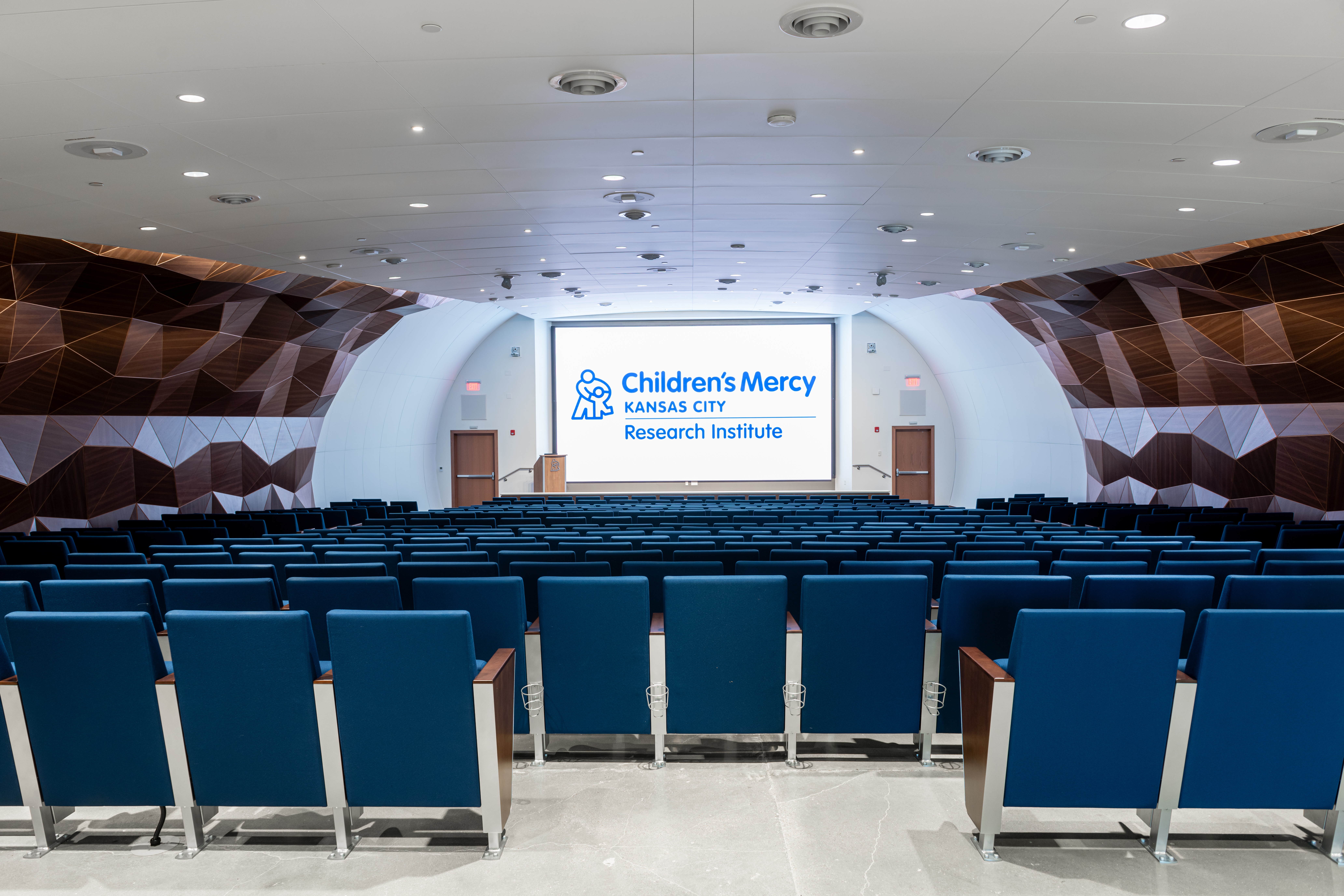 Inside the 370 seat CMRI Big Slick Auditorium. It is empty. On the screen at the front is a large slide that says Children's Mercy Kansas City Research Institute.