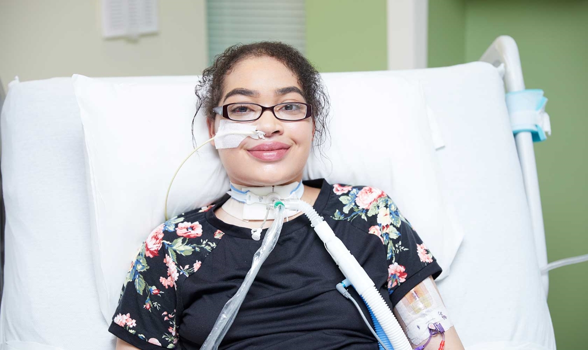 A teenage girl lays propped up in a hospital bed. She has a breathing tube coming out of her neck. She has black hair, is wearing glasses, and smiles at the camera.