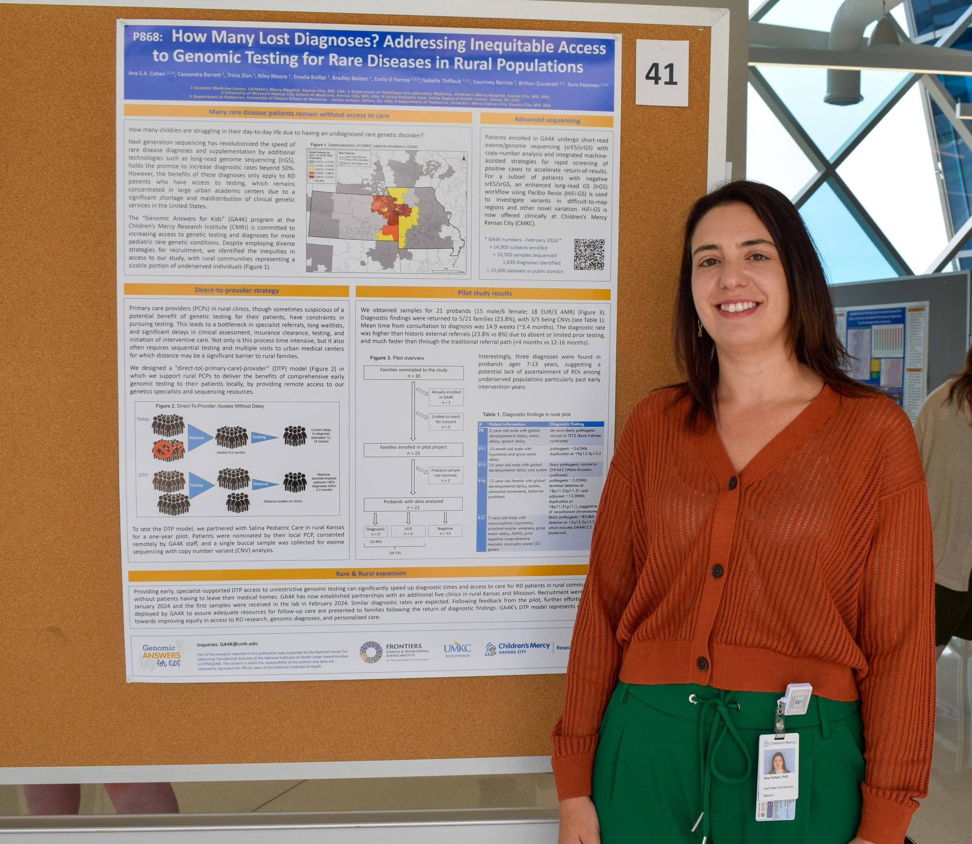 Dr. Ana Cohen, who has long dark hair and is wearing an burnt orange sweater, stands in front of her poster.