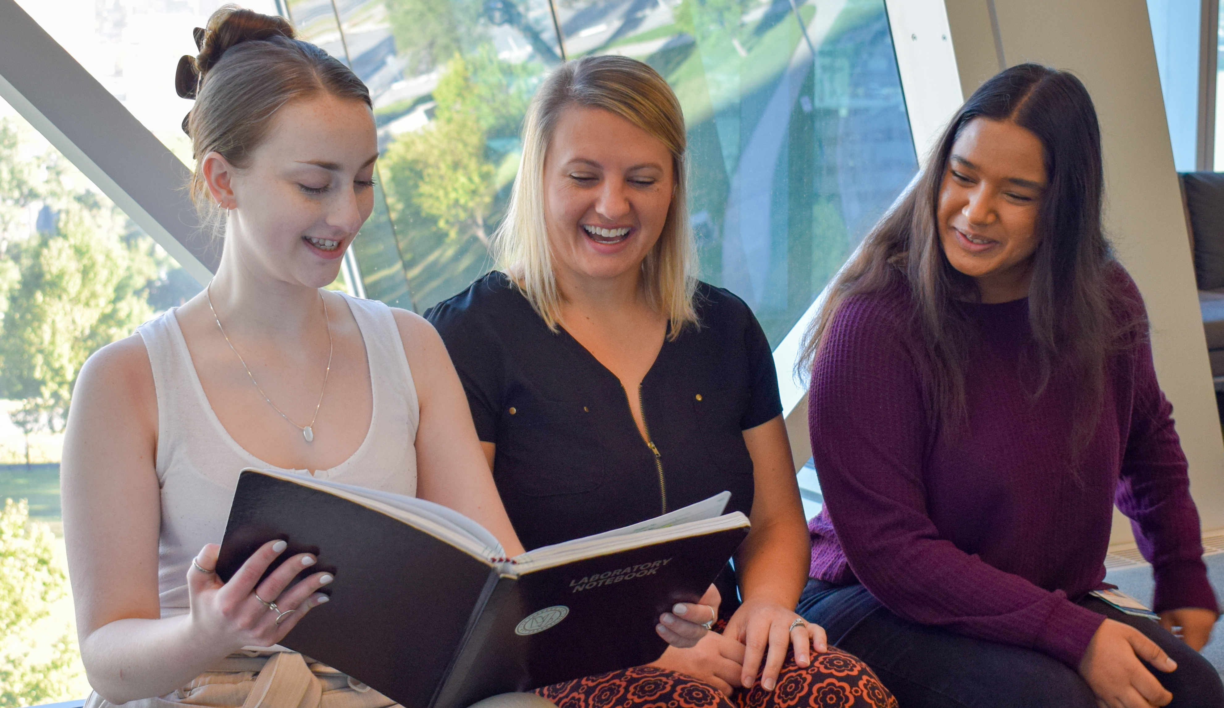 Three women sit on a bench looking at an open book. There is a large window behind them and it is daylight.
