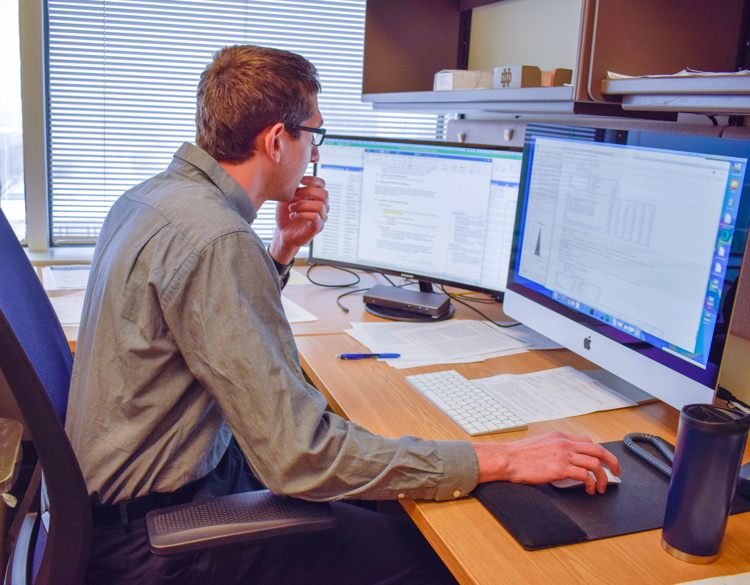 A man sits at a desk looking at two monitors. A cup, papers, and a pen are on the desk in front of him. He is wearing glasses.