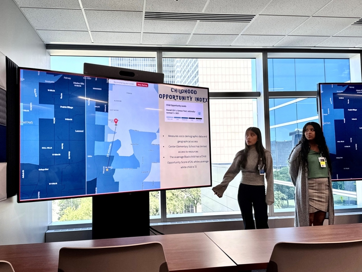 Two teenage women stand in a conference room, presenting. They stand next to a screen that has a map on it of various shades of blue and that says Childhood Opportunity Index at the top.