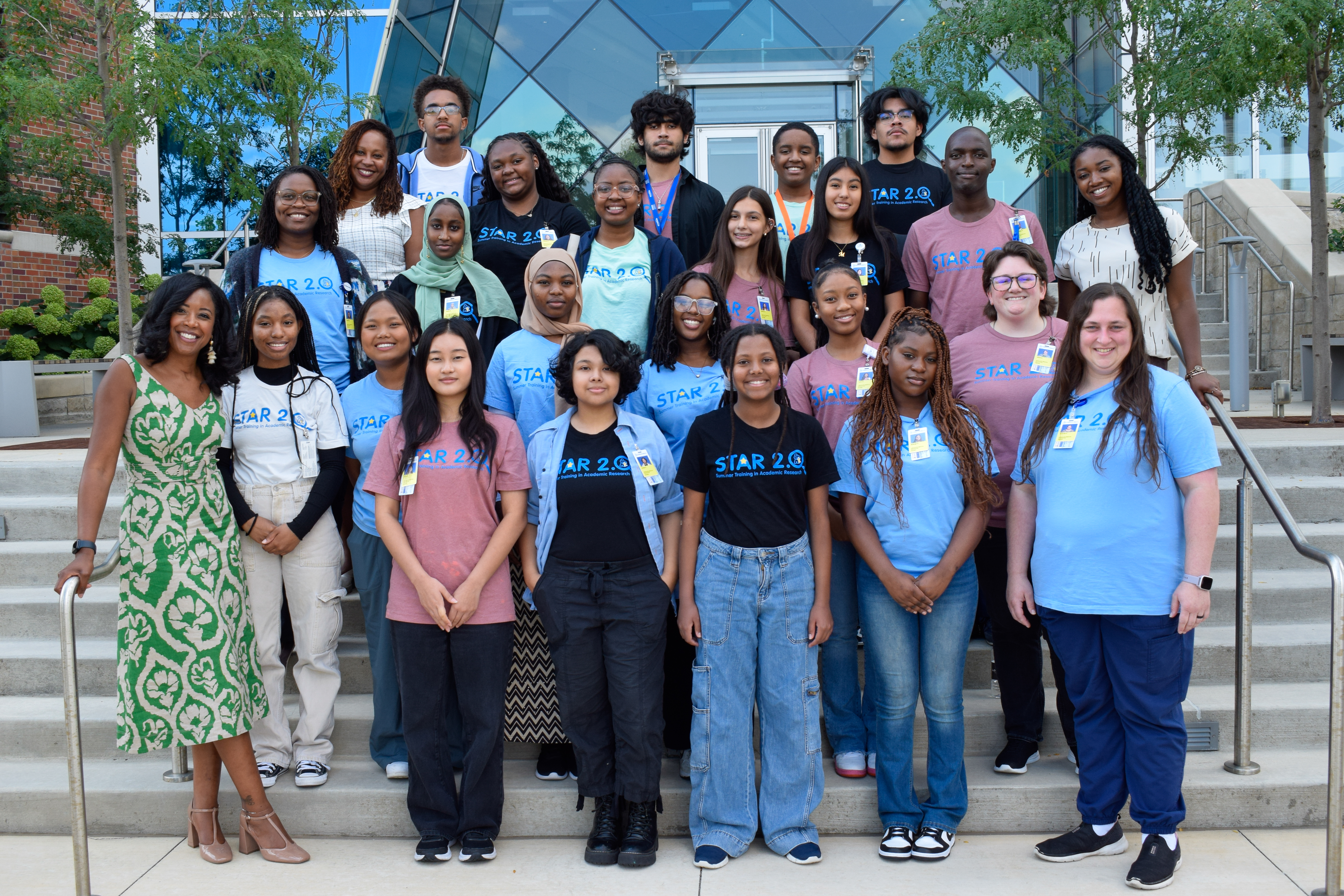 About 25 people, consisting of some adults but mostly high school students stand outside, posing on steps.