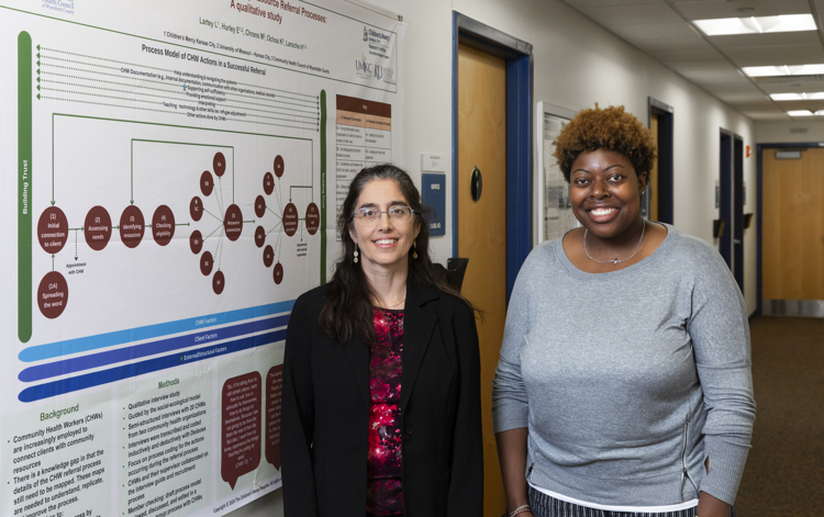 Two women stand next to each other in a hallway. On the wall on the left is a large research poster of the CHW referral process.