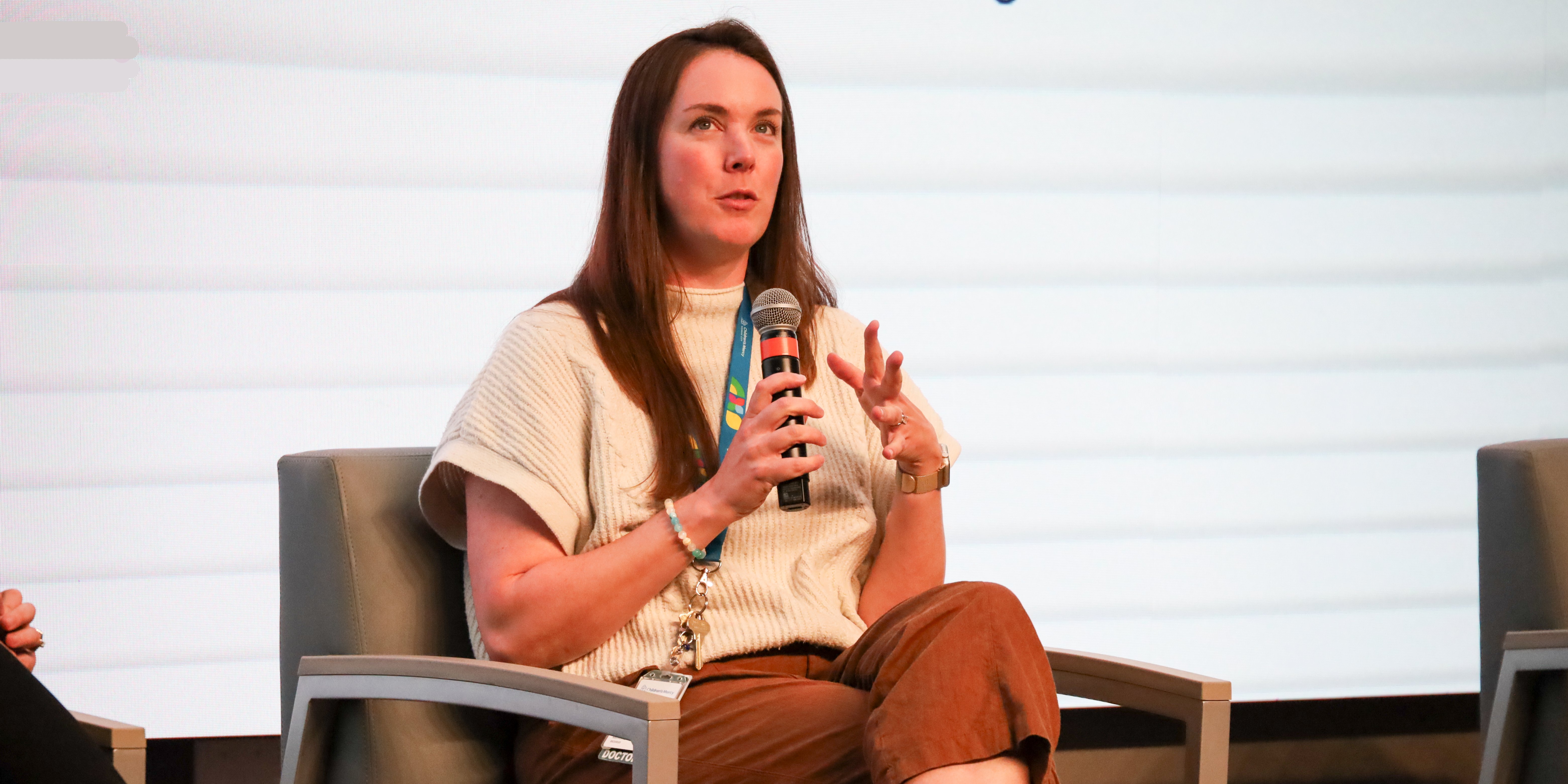 A woman with long brown hair, wearing a cream colored shirt and brown paints sits on a chair on a stage, holding a microphone, talking and gesturing with her hand.