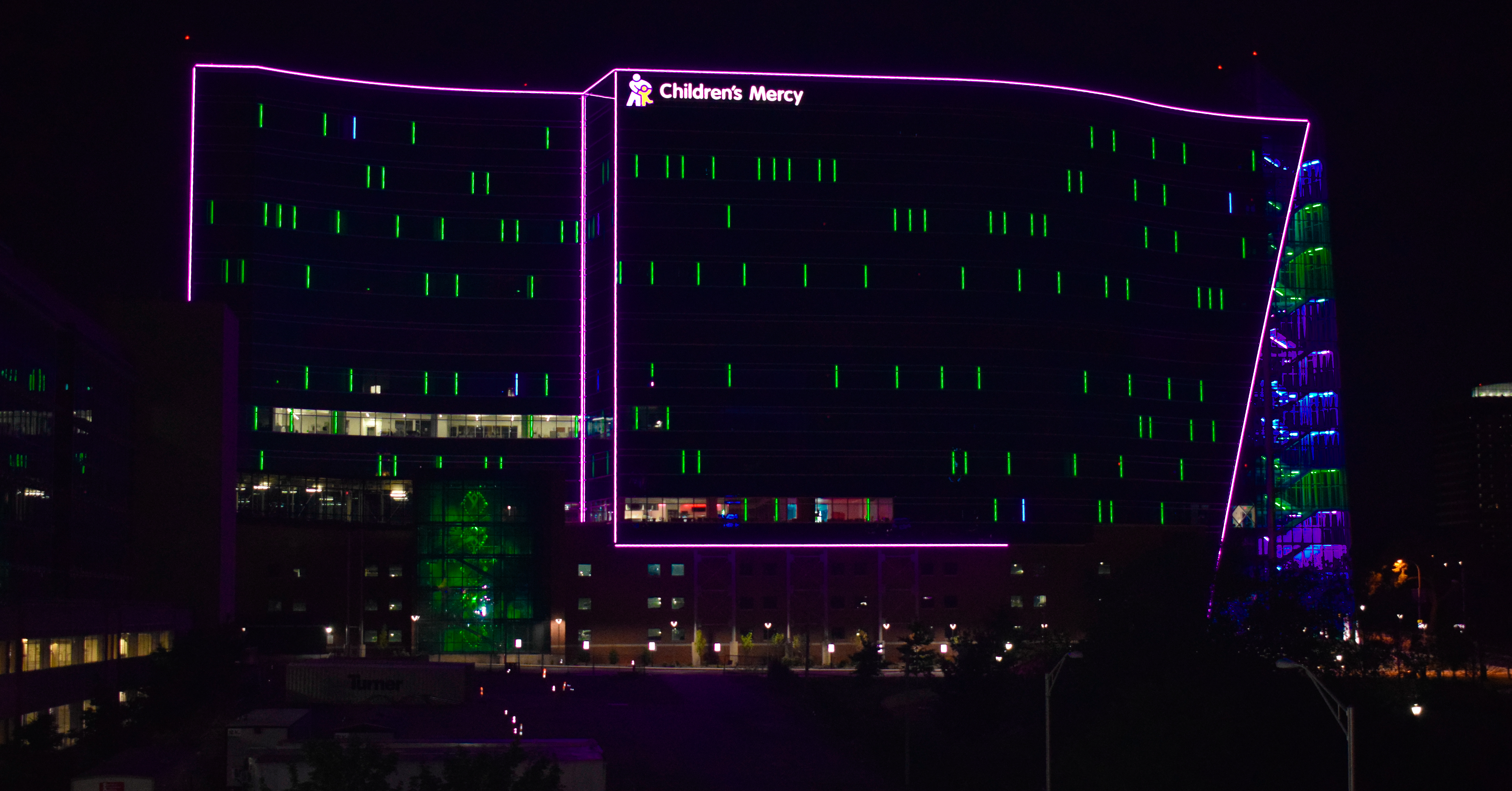 A night time photo of the outside of the Children's Mercy Research Institute building. It's overall dark except for blue lights along the outline and some blue lights inside the glass windows. 