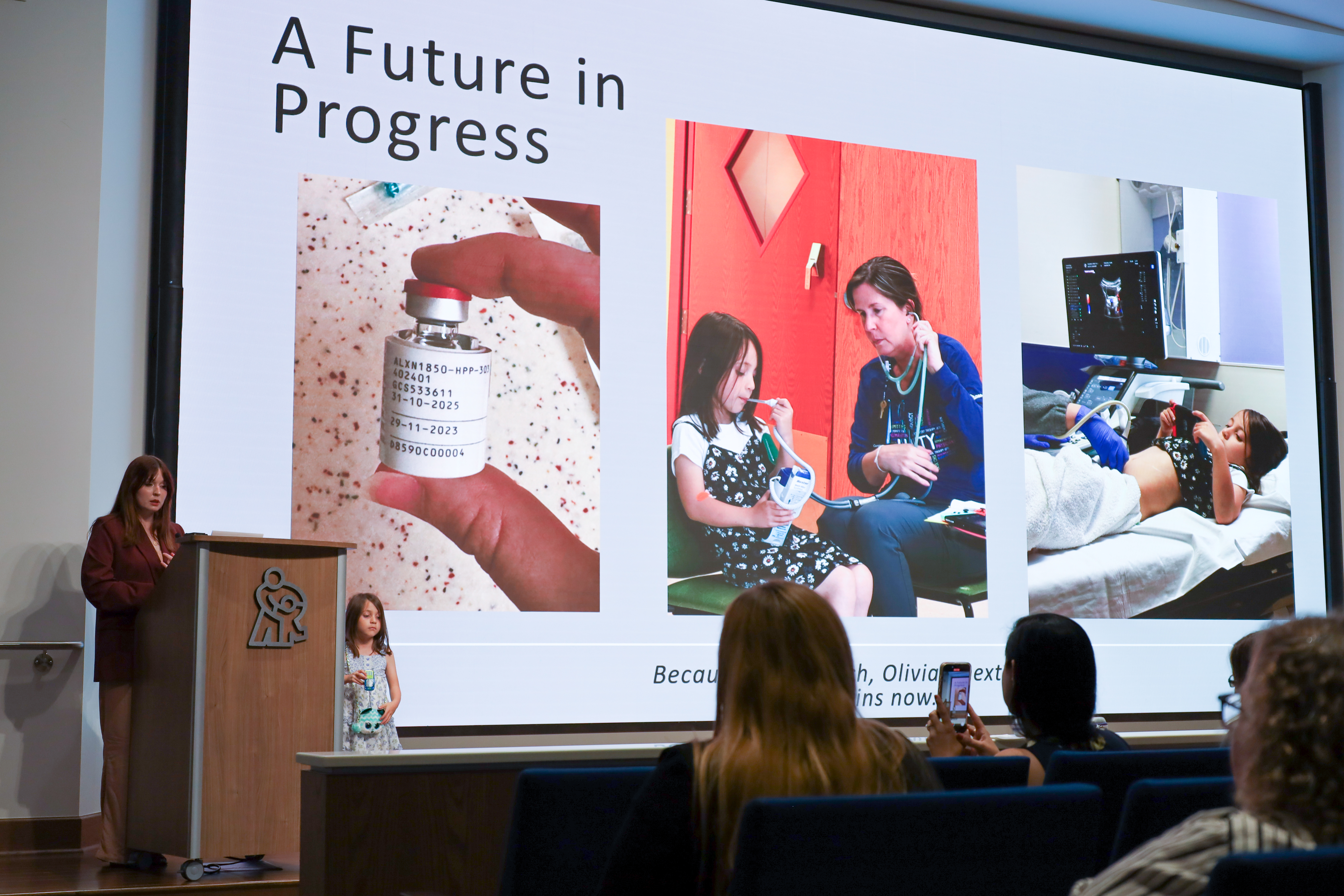 A woman and a young girl stand on stage at a lectern. Someone in the audience takes a photo on their cell phone. The large screen behind them has photos and says "A Future in Progress".