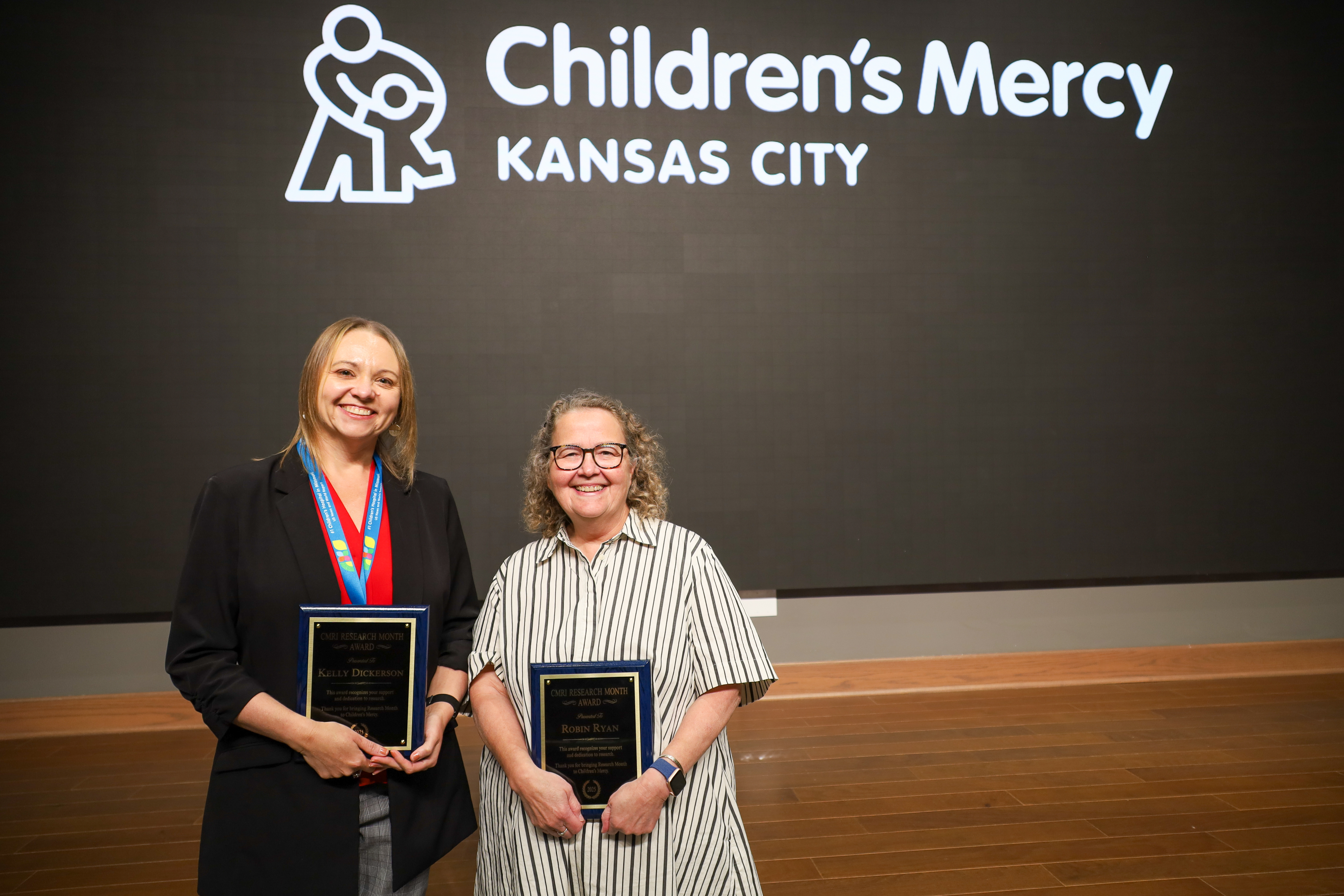Two women stand next to each other holding awards they received. 